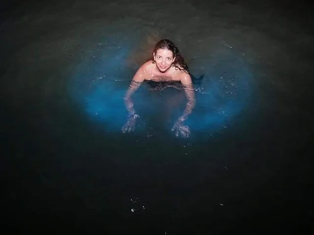 Une femme nage dans l'obscurité, entourée d'une eau bleue bioluminescente et luisante.
